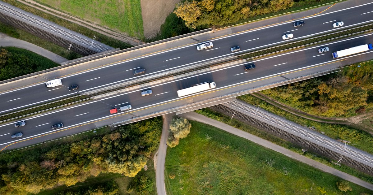 Luchtfoto van een snelweg met personenauto's en vrachtwagens in beide richtingen, met een viaduct en een spoorlijn tussen groene velden