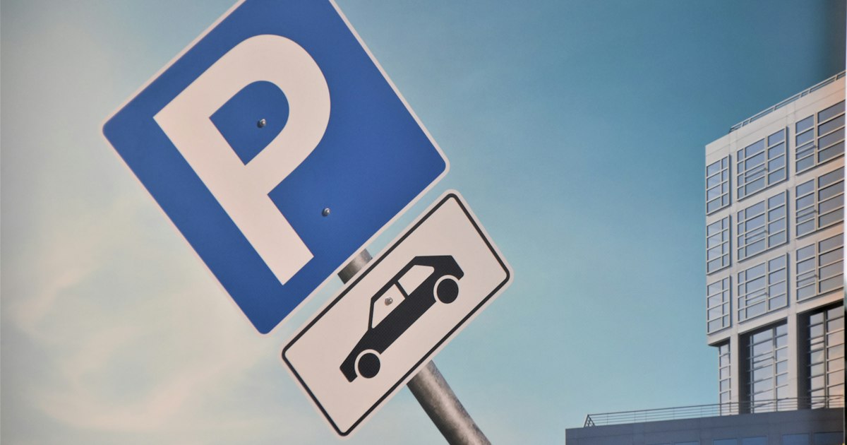 Blauw parkeerbord met een witte P en een onderbord met een auto, voor een kantoorgebouw tegen een blauwe lucht